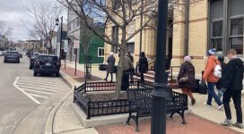 A group of people is walking in downtown Oconomowoc with two- and three-story buildings, a bench, trees, lamp posts, a mural on the side of a building, and full on-street parking.