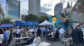 People are gathered around picnic tables in a parking lot surrounded by artistically decorated shipping containers with tall buildings in the background.