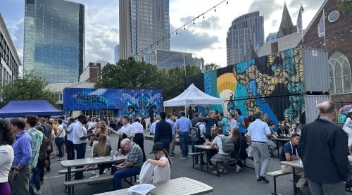 People are gathered around picnic tables in a parking lot surrounded by artistically decorated shipping containers with tall buildings in the background.